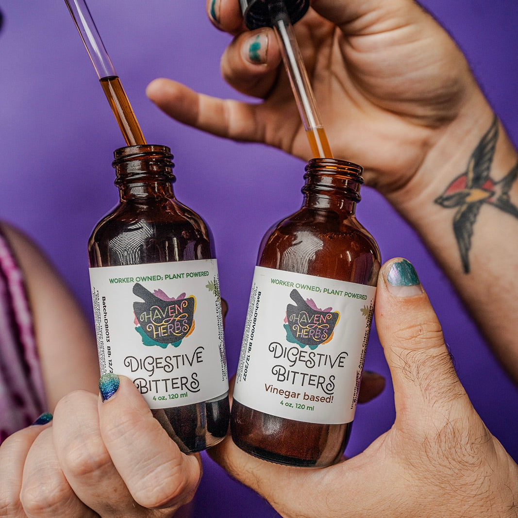 2 sets of hands pulling droppers from bottles of Digestive Bitters, on a purple background